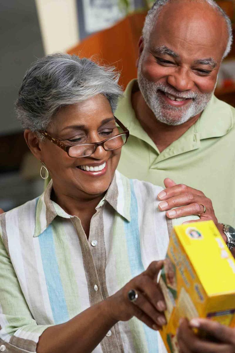 an older woman wearing glasses reads from the side of a box as her husband touches her shoulder